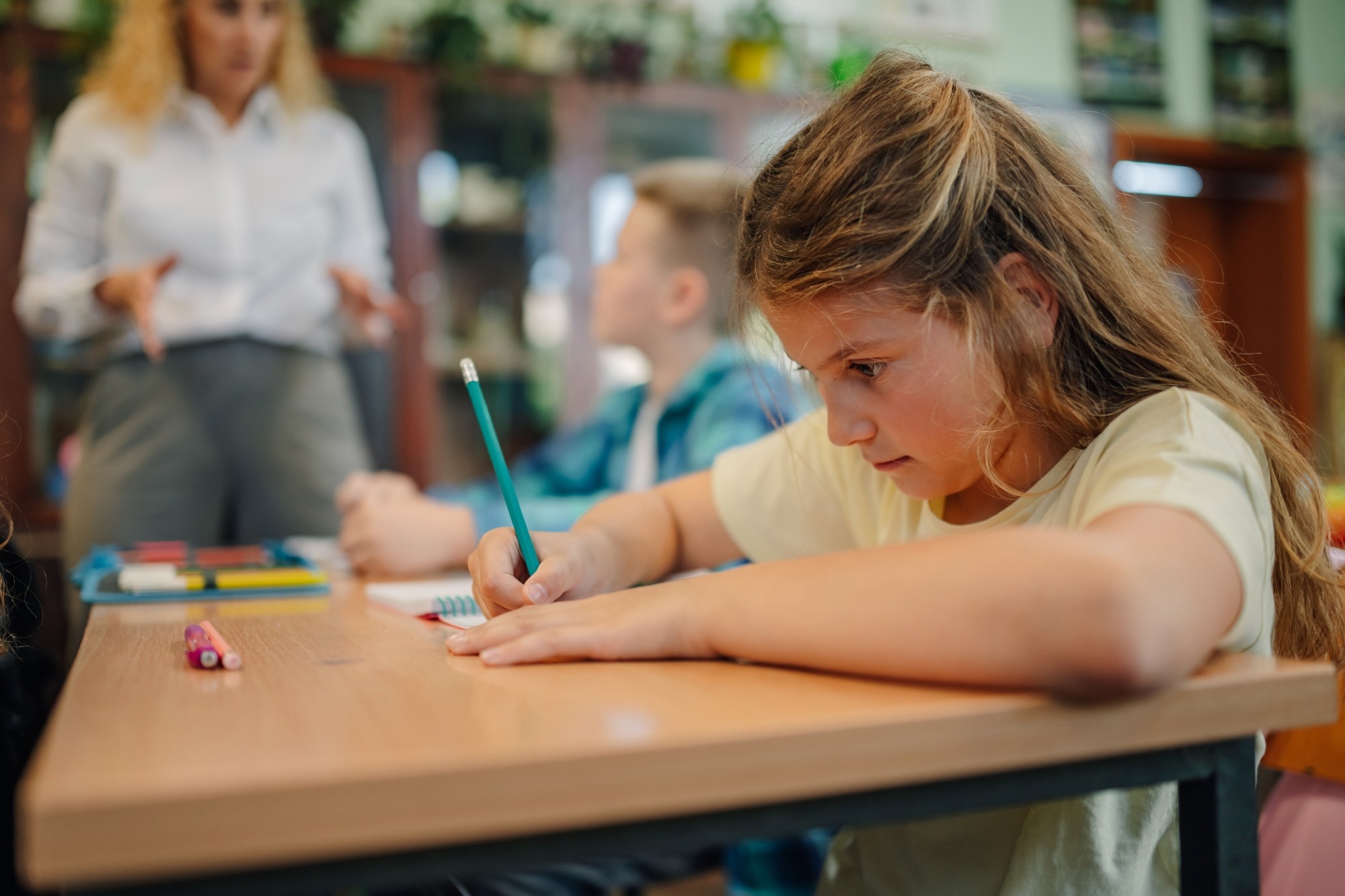 Une &eacute;coli&egrave;re &eacute;crivant sur un cahier avec un crayon pendant les cours, tandis que l'enseignant explique la le&ccedil;on aux &eacute;l&egrave;ves du primaire