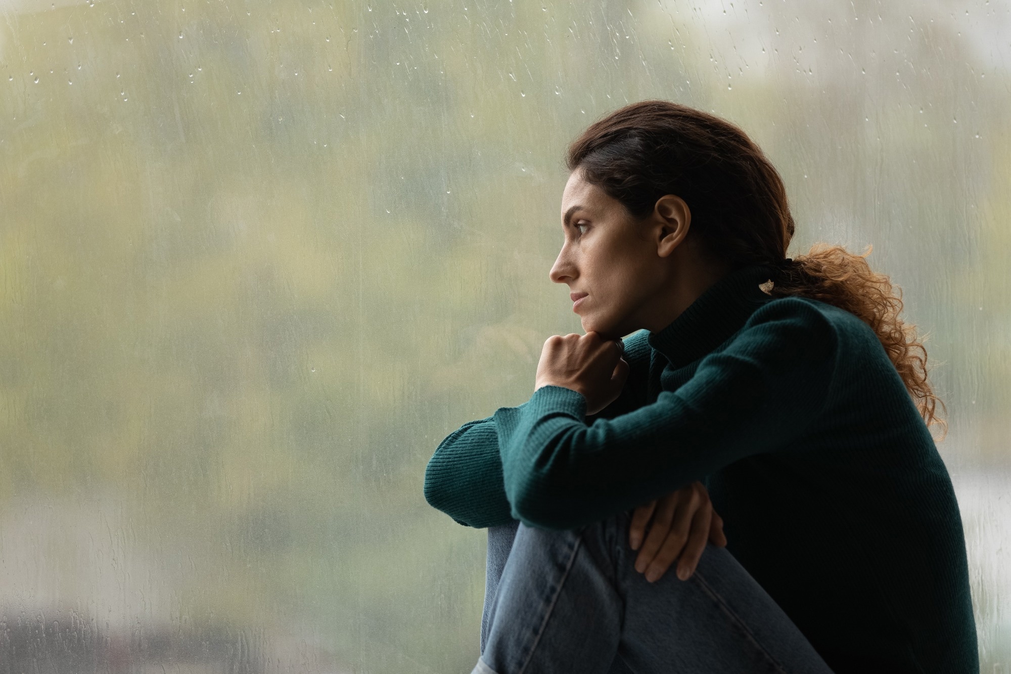 Vue lat&eacute;rale d'une femme r&eacute;fl&eacute;chie et frustr&eacute;e regardant par la fen&ecirc;tre pluvieuse seule &agrave; distance, perdue dans ses pens&eacute;es